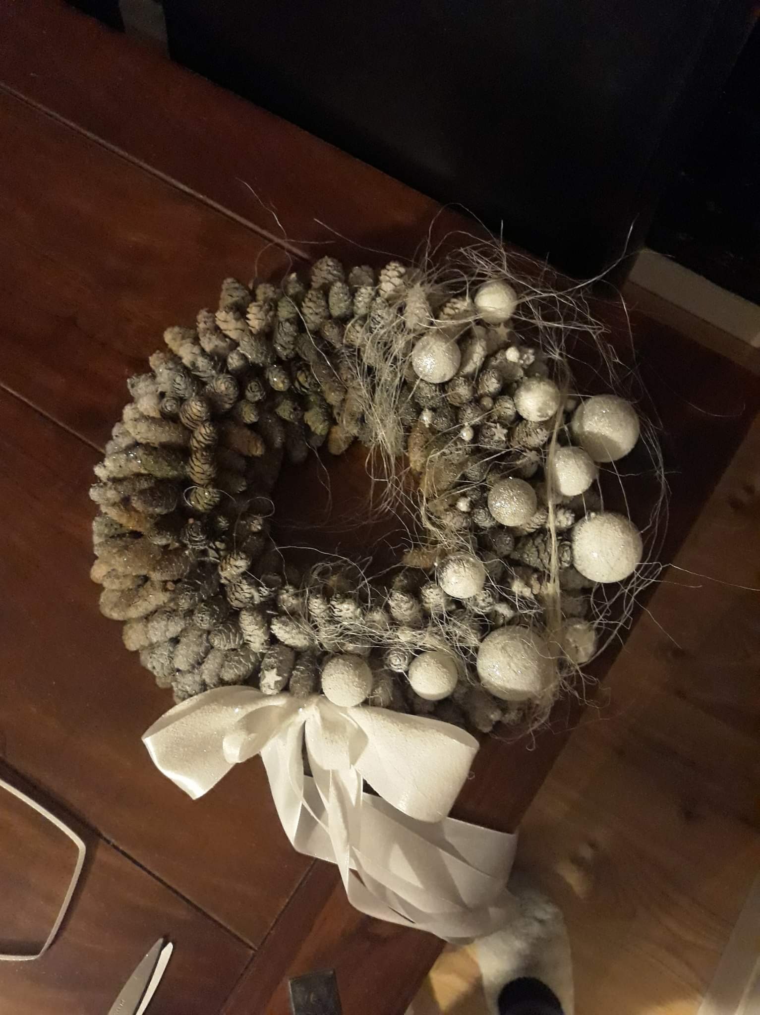 Wieniec z szyszek i białych bombek, ozdobiony błyszczącą wstążką, leżący na drewnianym stole.