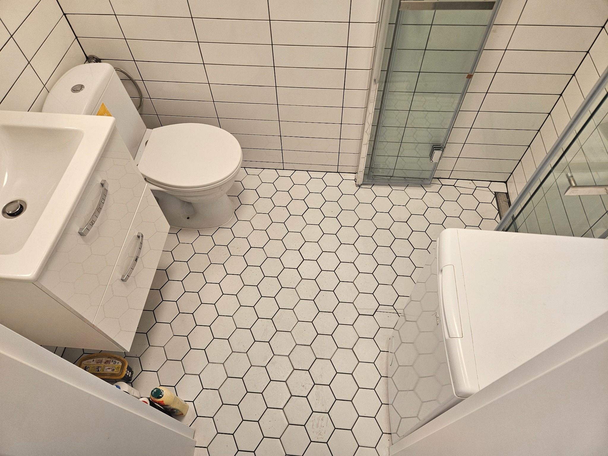 Widok z góry na wyremontowaną łazienkę z białą armaturą, heksagonalnymi płytkami podłogowymi i prostokątnymi płytkami ściennymi z czarną fugą, z widoczną pralką i kabiną prysznicową.