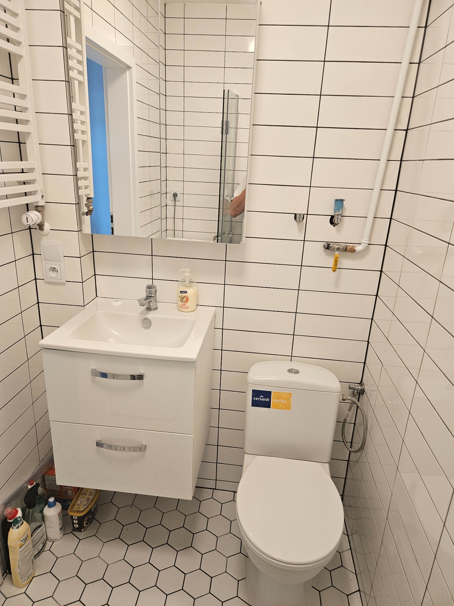 Biała łazienka z heksagonalną podłogą, wisząca szafka z umywalką, toaleta Cersanit Merida i grzejnik drabinkowy, płytki ścienne z czarną fugą.