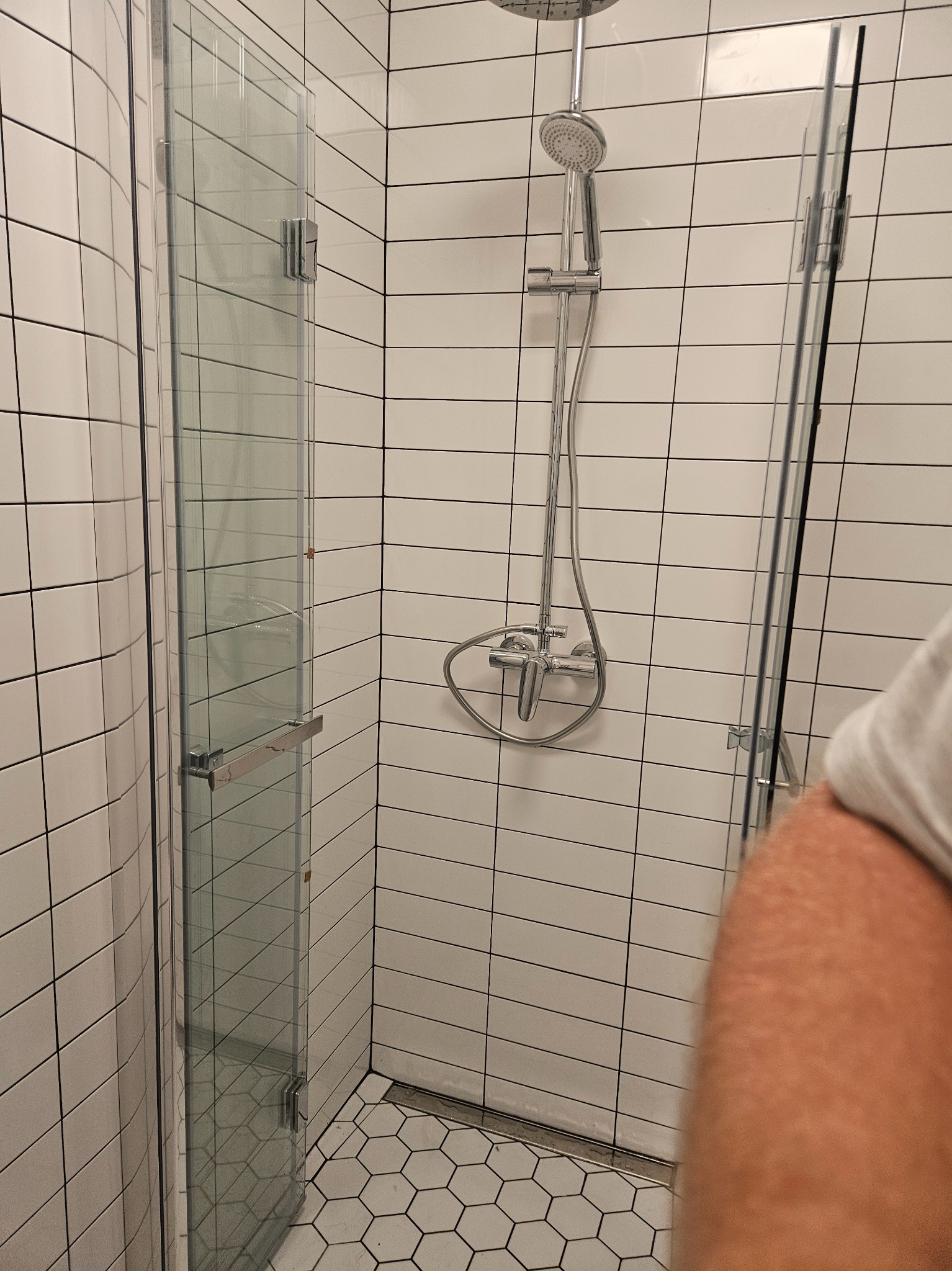 Kabina prysznicowa z chromowaną armaturą i białymi, prostokątnymi płytkami na ścianach, sześciokątne płytki podłogowe, widoczny fragment ciała osoby.