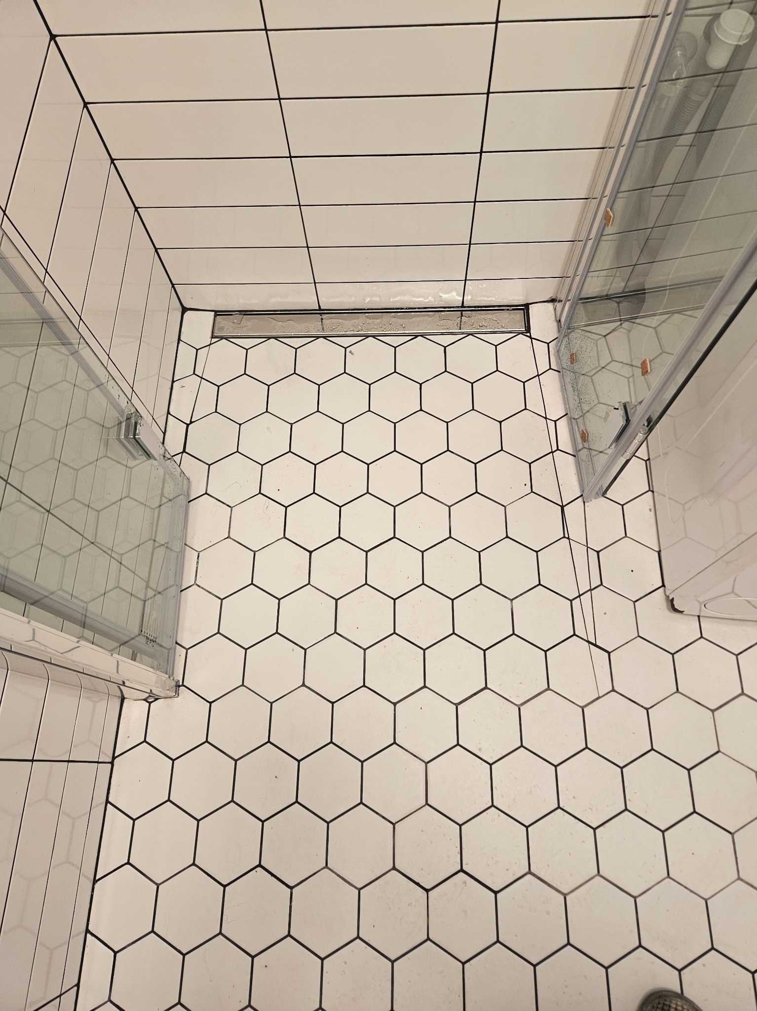 Biała łazienka z heksagonalnymi płytkami podłogowymi z czarną fugą i kabiną prysznicową z przezroczystego szkła, widok z góry na odpływ liniowy.