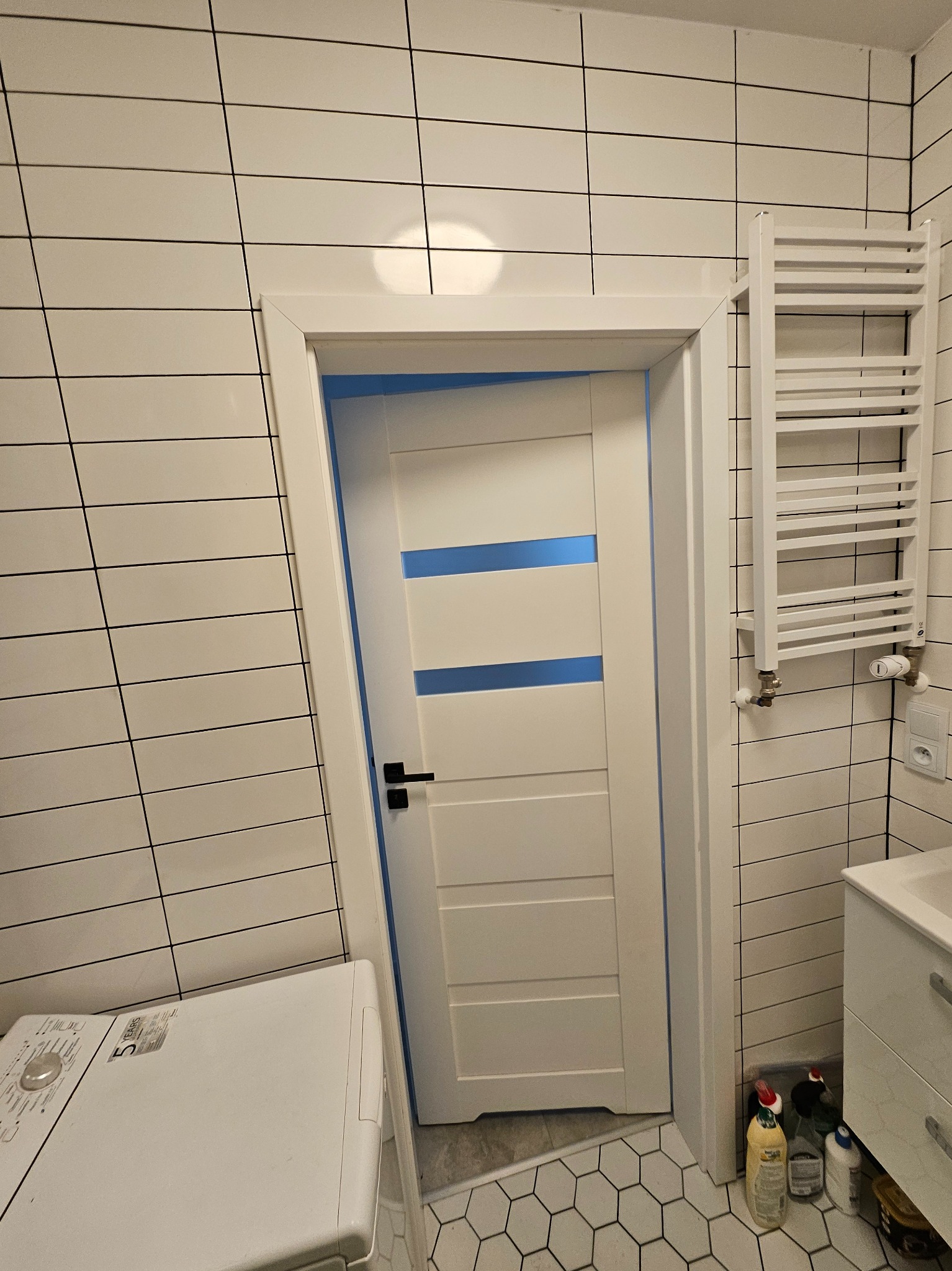 Biała łazienka z poziomymi płytkami, biała drabinka grzejnikowa, biała pralka i drzwi z niebieskimi wstawkami, sześciokątne płytki podłogowe.