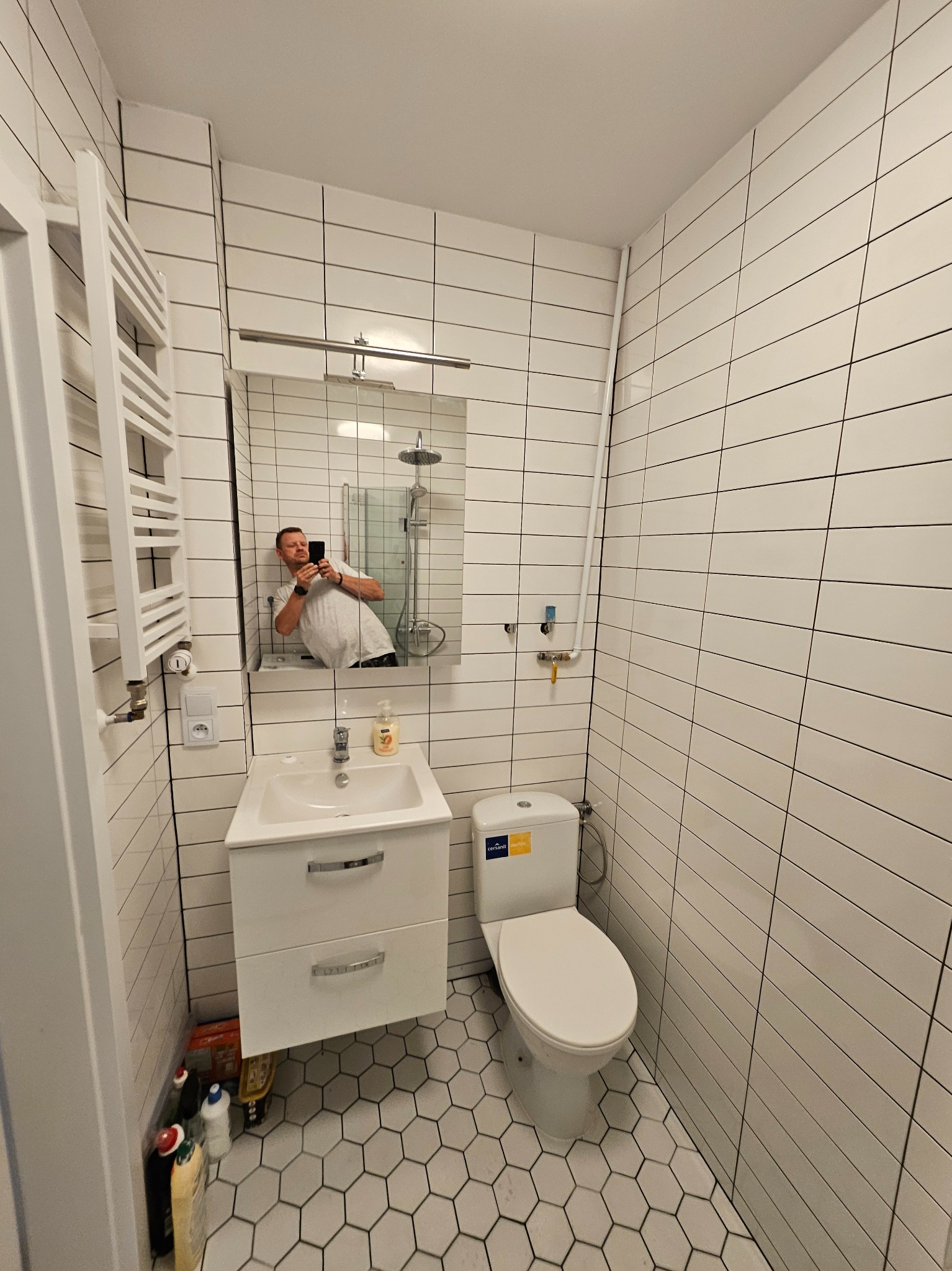 Nowoczesna łazienka z białymi, poziomymi płytkami, heksagonalną podłogą i odbiciem fotografa w lustrze nad umywalką.