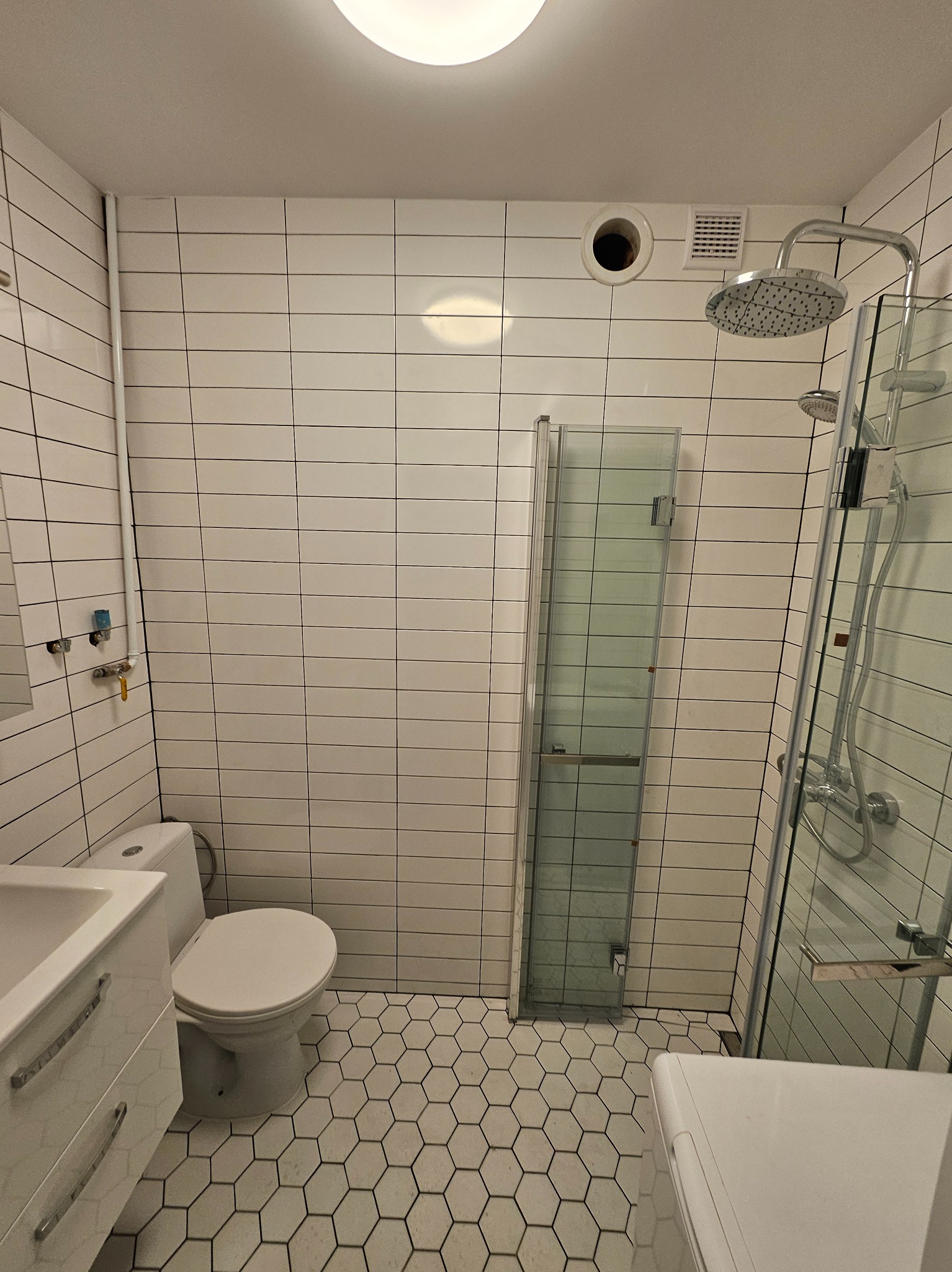 Biała łazienka z heksagonalną podłogą, prysznicem typu walk-in i umywalką z szafką, wykończona płytkami imitującymi cegłę.