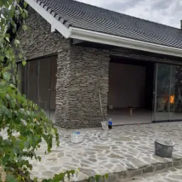 Budynek z elewacją z kamienia łupanego, z dużymi przesuwnymi oknami, na wykończonym kamiennym tarasie, widoczne narzędzia i materiały budowlane.