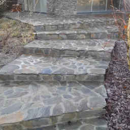 Kamienne schody prowadzące do domu z elewacją z kamienia łupanego i dużymi oknami.