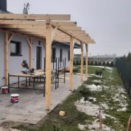 Montaż drewnianej pergoli przy szarym domu z widocznymi narzędziami, stołem roboczym, puszkami farby i resztkami śniegu na trawniku.