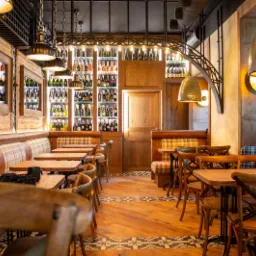 Przytulne wnętrze restauracji z drewnianymi stołami, krzesłami i kanapami w kratę, podłoga w jasnym drewnie z wzorzystym pasem, ściana z cegły i lustra, regał z butelkami podświetlony od tyłu...