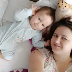 Uśmiechnięta kobieta robi selfie z leżącym obok niemowlęciem w błękitnym pajacyku z aplikacją śpiącego liska, na białej pościeli z fioletowymi elementami i pluszową zabawką w tle.