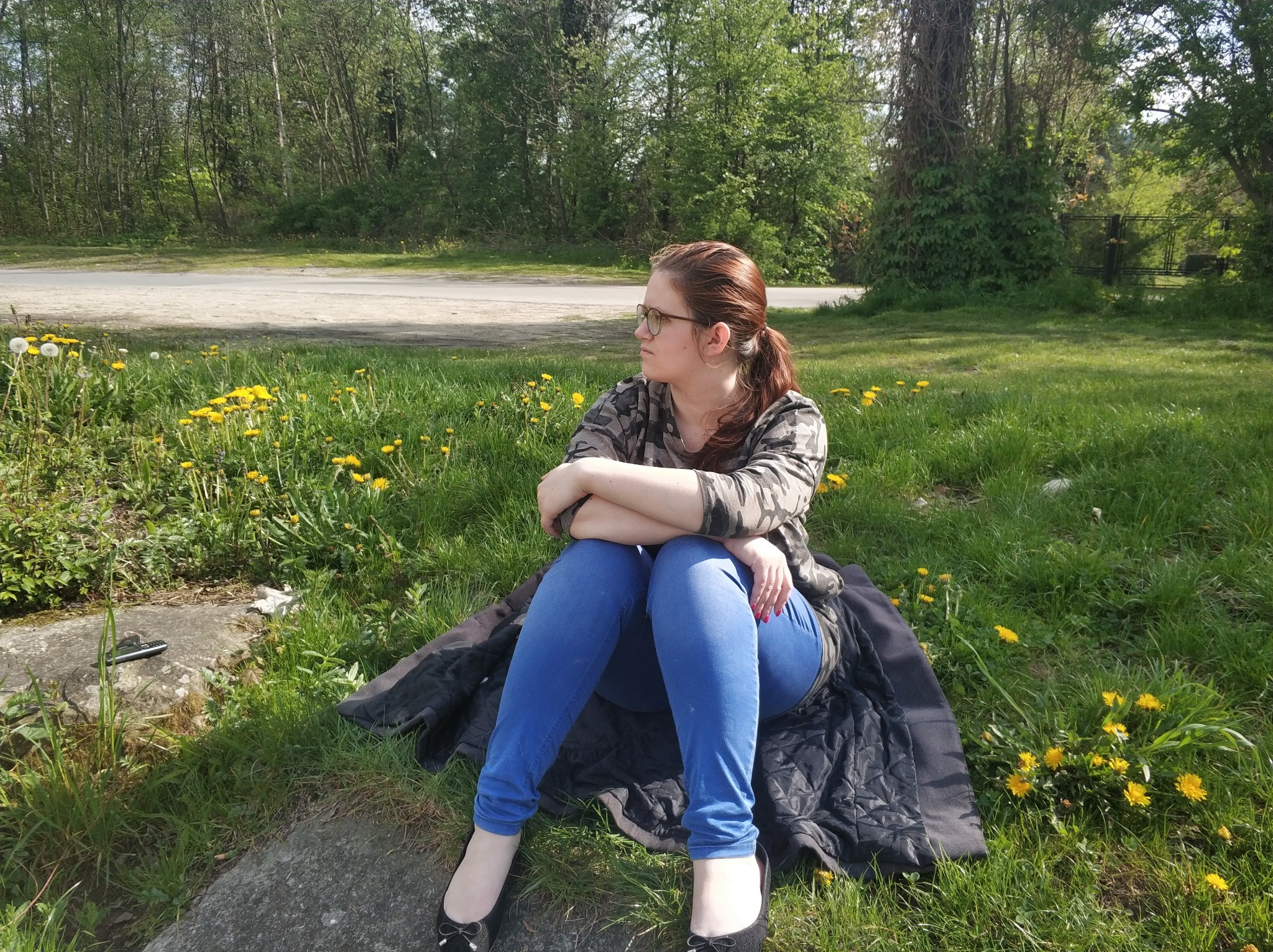 Kobieta w okularach siedzi na trawie w parku, ubrana w jeansy i bluzę moro, spoglądając w dal.