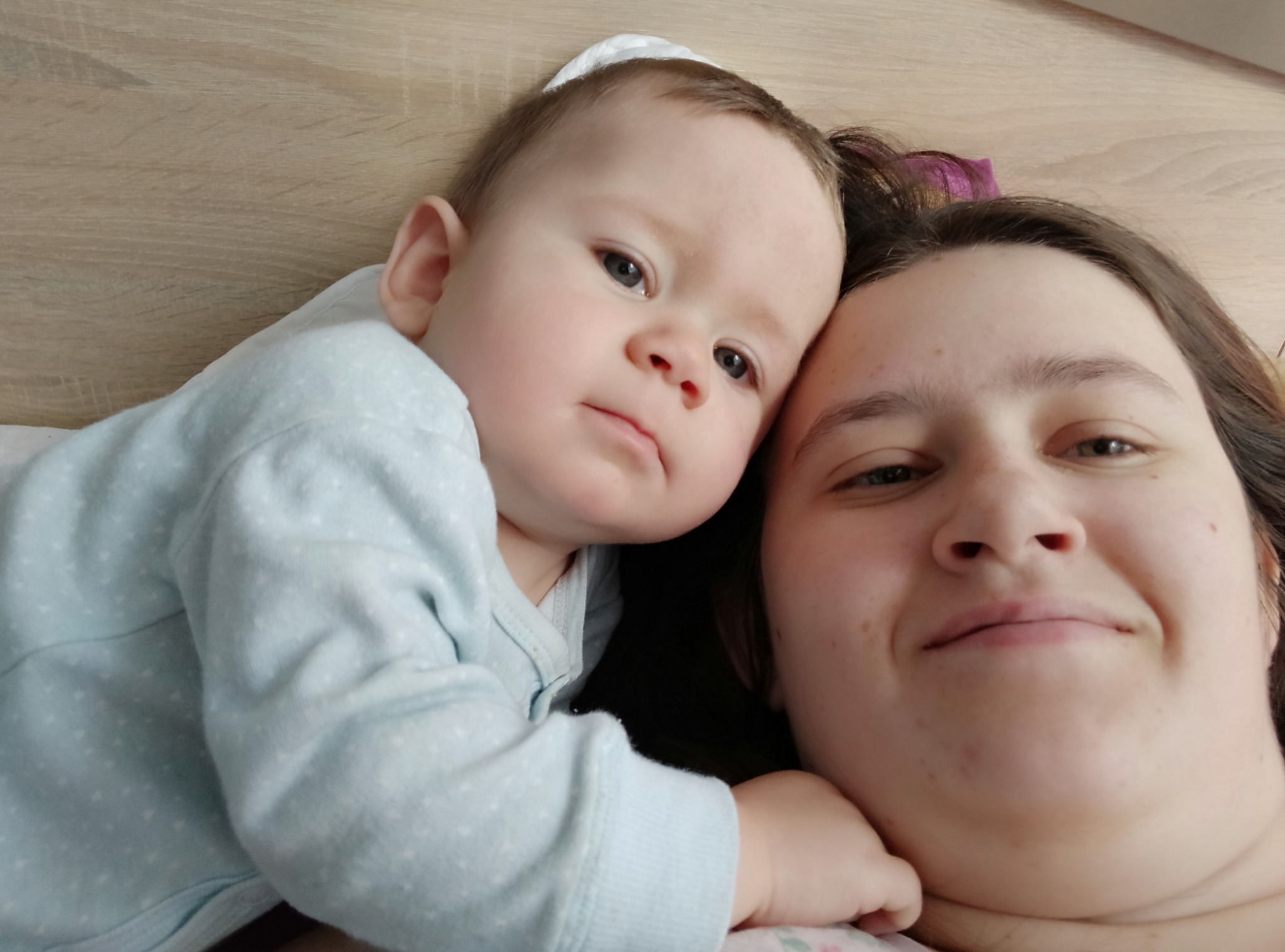 Uśmiechnięta kobieta z ciemnymi włosami i małe dziecko w jasnoniebieskim ubranku leżące obok siebie na jasnym tle.