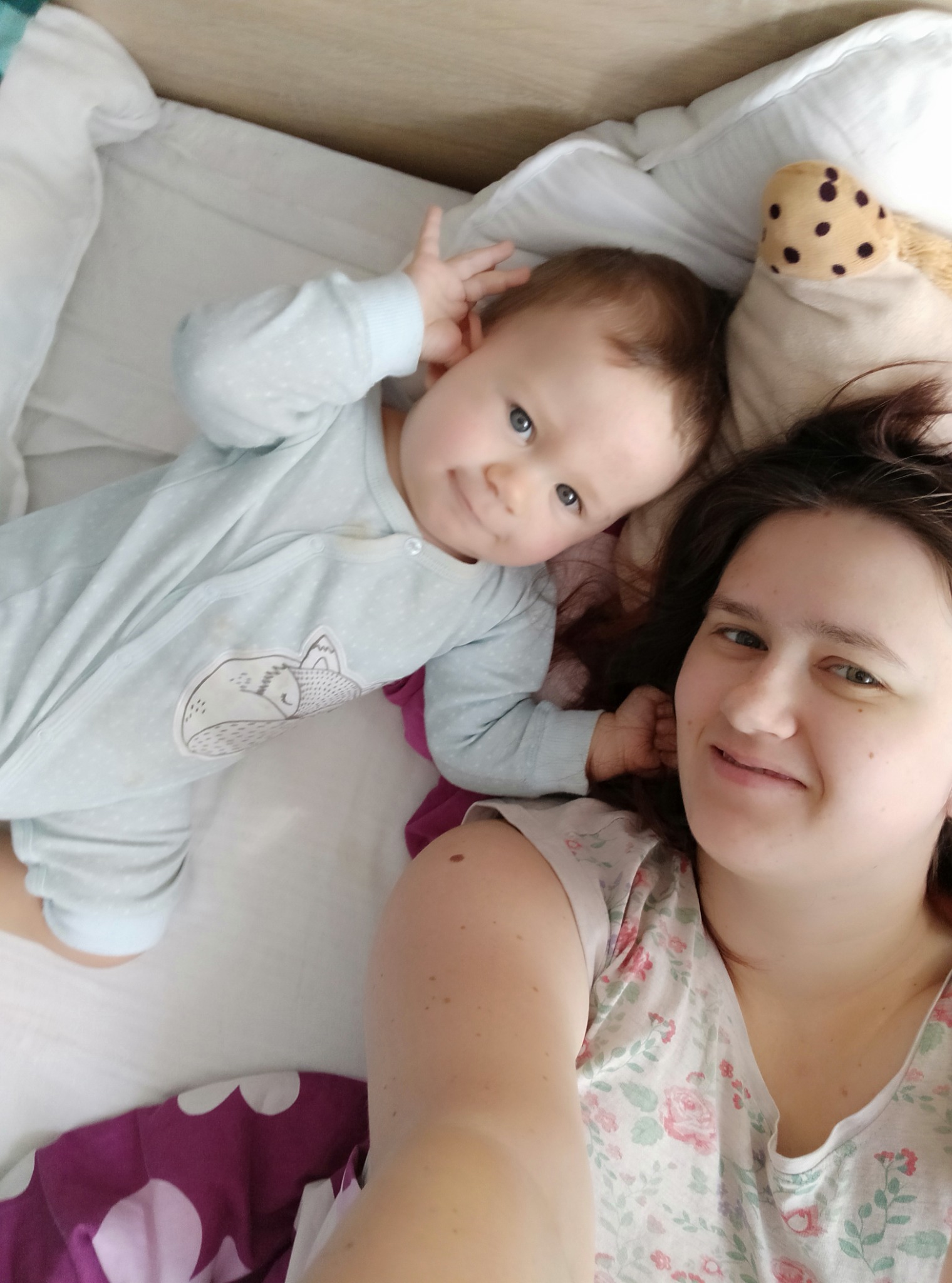 Uśmiechnięta kobieta robi selfie z leżącym obok niemowlęciem w błękitnym pajacyku z aplikacją śpiącego liska, na białej pościeli z fioletowymi elementami i pluszową zabawką w tle.