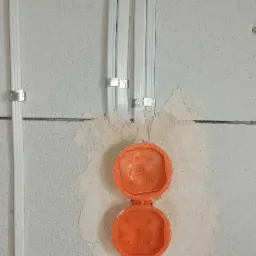 Białe rury elektryczne przymocowane metalowymi uchwytami do szarej ściany z betonu komórkowego, z otwartą, pomarańczową puszką elektryczną w ścianie.