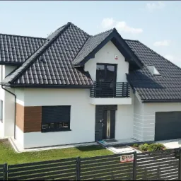 Nowoczesny dom jednorodzinny z ciemną dachówką, białymi ścianami i elementami dekoracyjnymi imitującymi drewno, widoczny z zewnątrz, z zadbanym trawnikiem i ogrodzeniem panelowym.