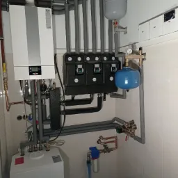 Nowoczesna kotłownia z piecem gazowym Wolf, rozdzielaczami z manometrami, zabezpieczeniami i filtrem wody, instalacja w pomieszczeniu wyłożonym białymi płytkami.