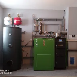 Kotłownia z kotłem na pellet, zbiornikiem akumulacyjnym Kospel i instalacją CO z zaworami i manometrami, widok z przodu na tle szarej ściany.