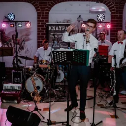 Wokalista w okularach i białej koszuli z muszką, śpiewa podczas występu, za nim perkusista, gitarzysta i basista, na scenie akordeon i saksofon, w tle ceglana ściana i dekoracja z motywem jelenia.