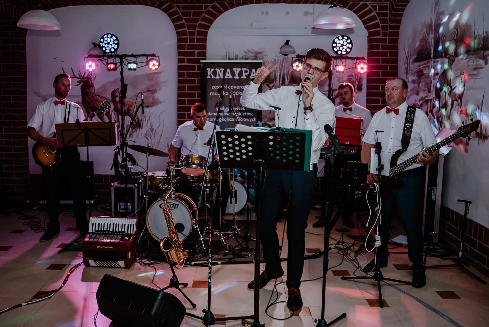 Wokalista w okularach i białej koszuli z muszką, śpiewa podczas występu, za nim perkusista, gitarzysta i basista, na scenie akordeon i saksofon, w tle ceglana ściana i dekoracja z motywem jelenia.