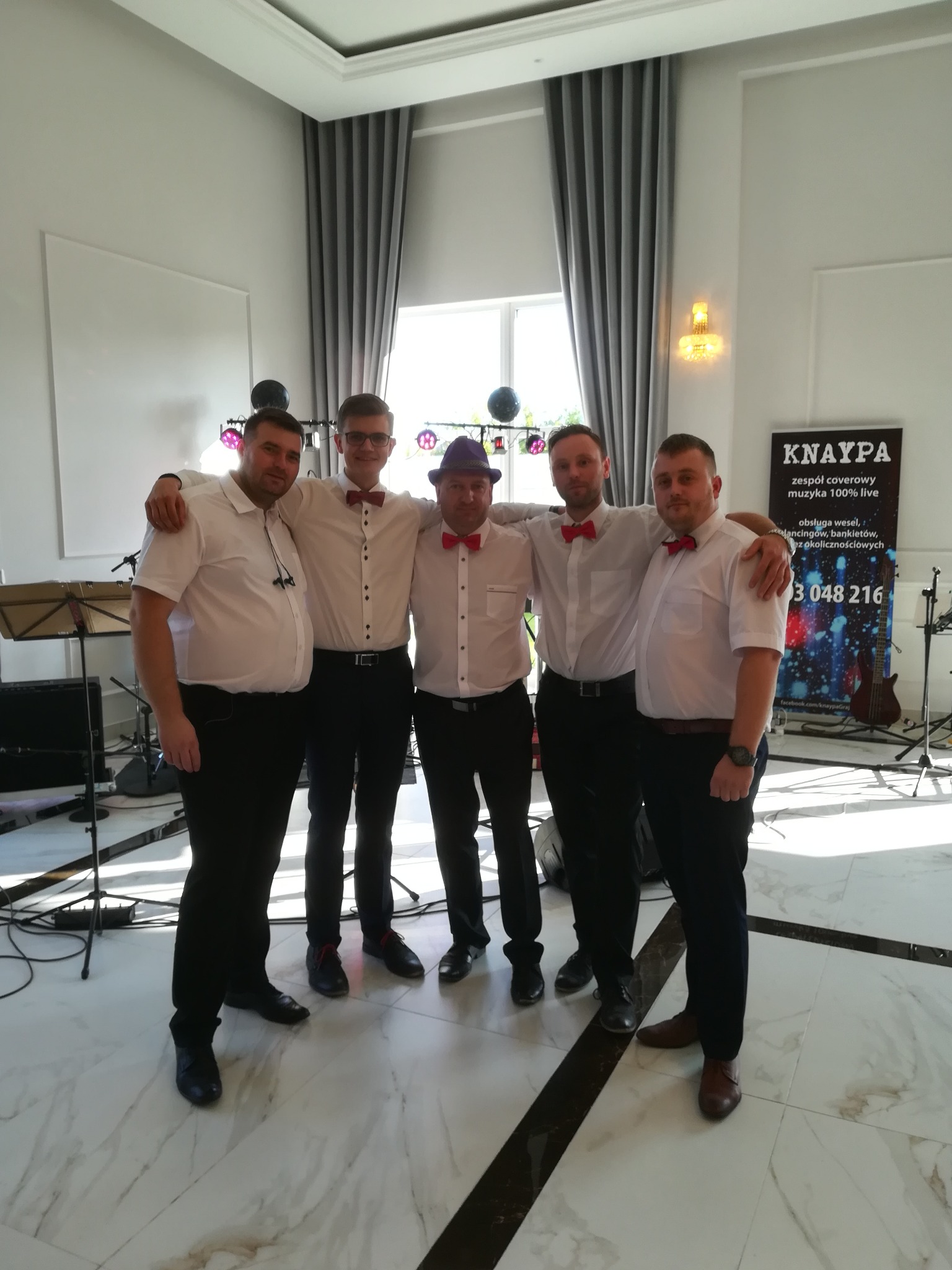 Pięciu mężczyzn w koszulach i muchach, stojących ramię w ramię na marmurowej podłodze, za nimi sprzęt muzyczny i baner reklamowy z nazwą zespołu 'KNAYPA', w tle okno z szarymi zasłonami.