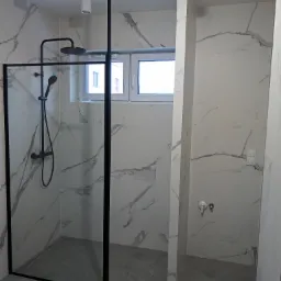 Kabina prysznicowa typu walk-in z czarną ramą i deszczownicą, płytki imitujące marmur na ścianach, szare płytki ułożone w jodełkę na podłodze, widoczne okno i oświetlenie sufitowe.