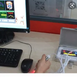 Dłoń trzyma elektrodę urządzenia Quantum Resonance Magnetic Analyzer, obok otwarty metalowy futerał, ekran komputera z wykresami i schematem ciała.