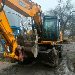 Żółta koparka kołowa JCB JS200 z łyżką pełną błota na placu budowy z widocznymi zbiornikami i stertą cegieł w tle.