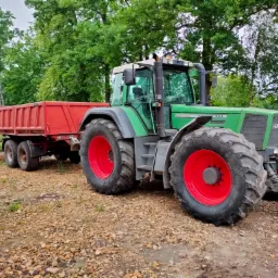 Zielony traktor Fendt z czerwoną przyczepą na żwirowym podłożu, otoczony zielenią drzew.