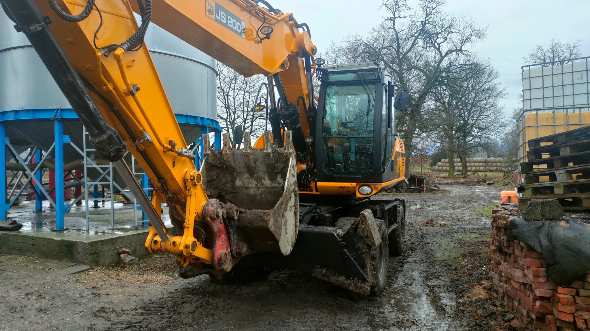 Żółta koparka kołowa JCB JS200 z łyżką pełną błota na placu budowy z widocznymi zbiornikami i stertą cegieł w tle.