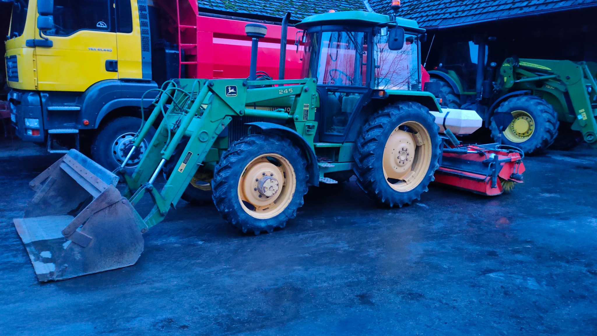 Zielony traktor John Deere 245 z łyżką i szczotką na tle żółtej ciężarówki MAN i czerwonej przyczepy, z drugim traktorem widocznym w tle.