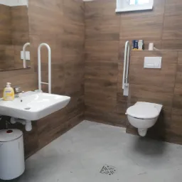 Wykończona łazienka z brązowymi płytkami imitującymi drewno, przystosowana dla osób niepełnosprawnych z umywalką, toaletą podwieszaną i uchwytami pomocniczymi. Widoczny podgrzewacz wody Atlantic...