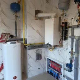 Wnętrze kotłowni z białym piecem gazowym, zbiornikiem ciepłej wody Galmet i rozdzielaczem ogrzewania podłogowego, widoczne rury w izolacji i manometry na marmurowych płytkach.