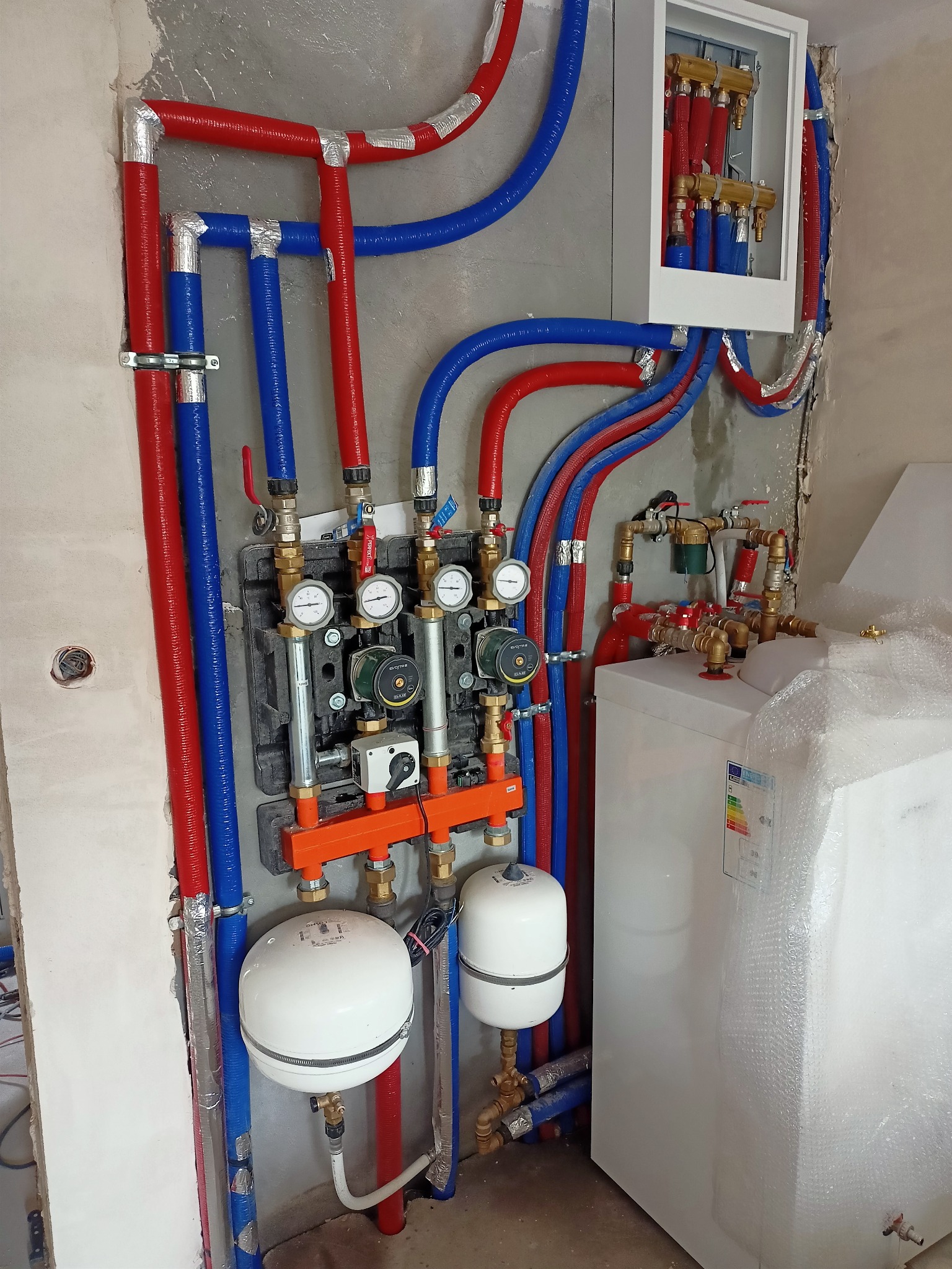 Świeżo zamontowana instalacja centralnego ogrzewania z piecem, zbiornikiem wyrównawczym i rozdzielaczem, rury w izolacji w kolorach niebieskim i czerwonym, manometry i zawory, część pieca owinięta...