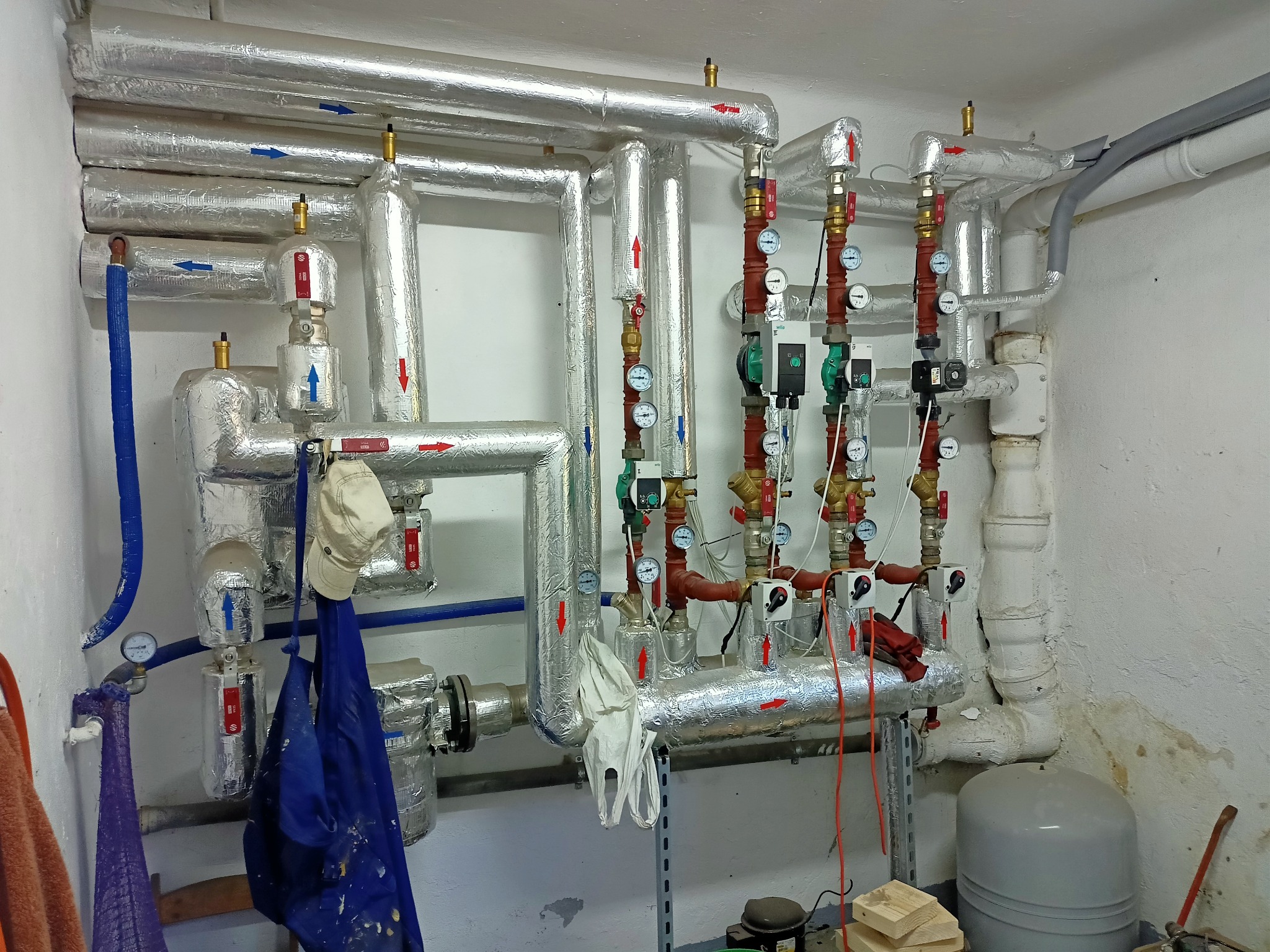Złożona instalacja grzewcza z rurami zaizolowanymi folią aluminiową, manometry, zawory, wiszące ubrania robocze i czapka, widoczne elementy kotłowni