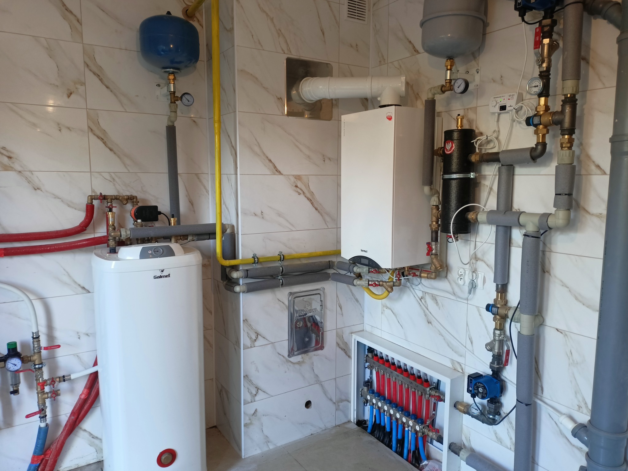 Wnętrze kotłowni z białym piecem gazowym, zbiornikiem ciepłej wody Galmet i rozdzielaczem ogrzewania podłogowego, widoczne rury w izolacji i manometry na marmurowych płytkach.