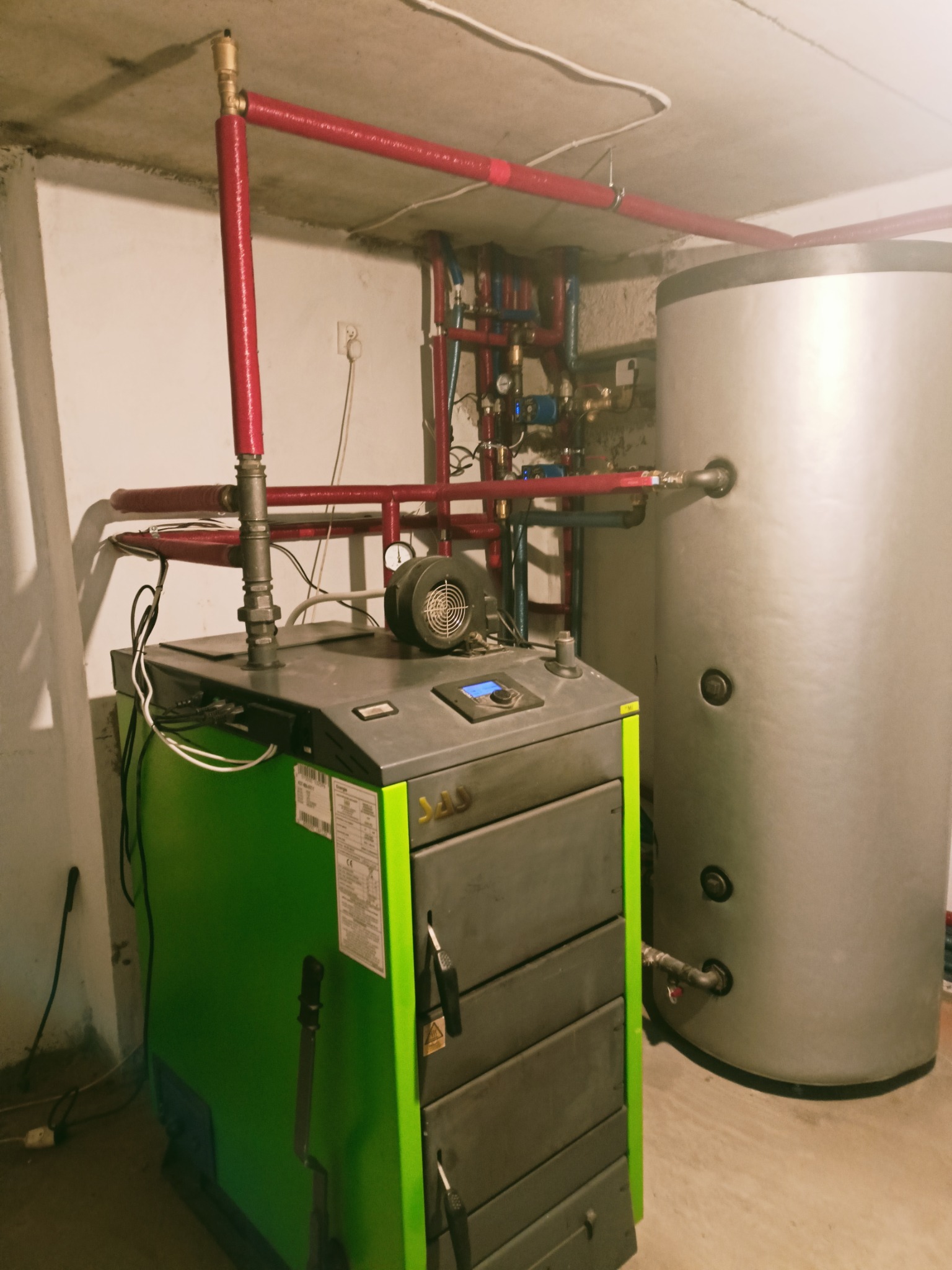 Nowoczesny piec na paliwo stałe, z zielonym wykończeniem, podłączony do instalacji centralnego ogrzewania z czerwonymi rurami i zasobnikiem ciepłej wody użytkowej w kotłowni.