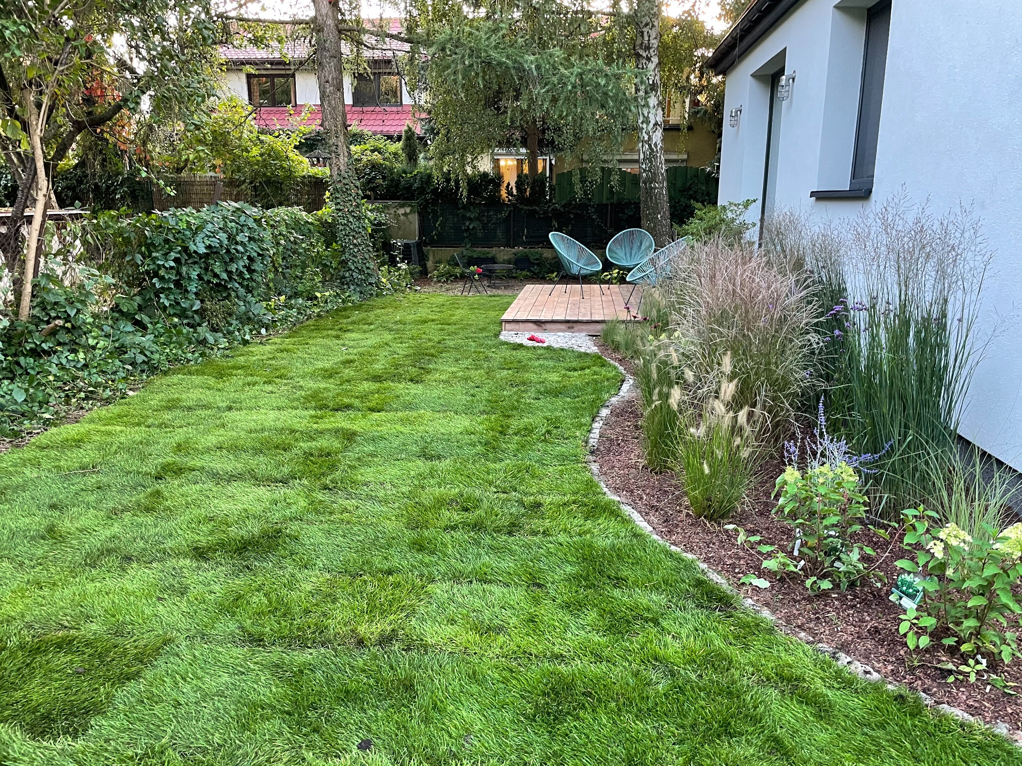 Świeżo założony trawnik z rolki, obrzeże z kory, ozdobne trawy i niskie krzewy przy białej ścianie domu, w tle drewniany taras z turkusowymi krzesłami.