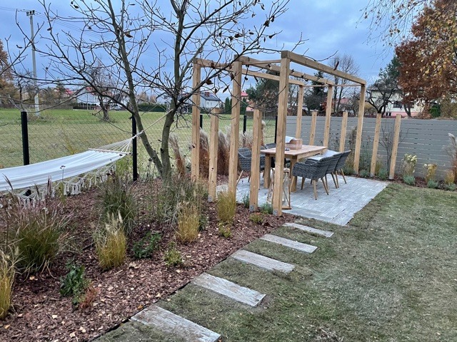 Ogród z drewnianą pergolą nad stołem z krzesłami na kostce brukowej, ścieżka z płyt, trawa, ozdobne trawy i hamak zawieszony na drzewie.