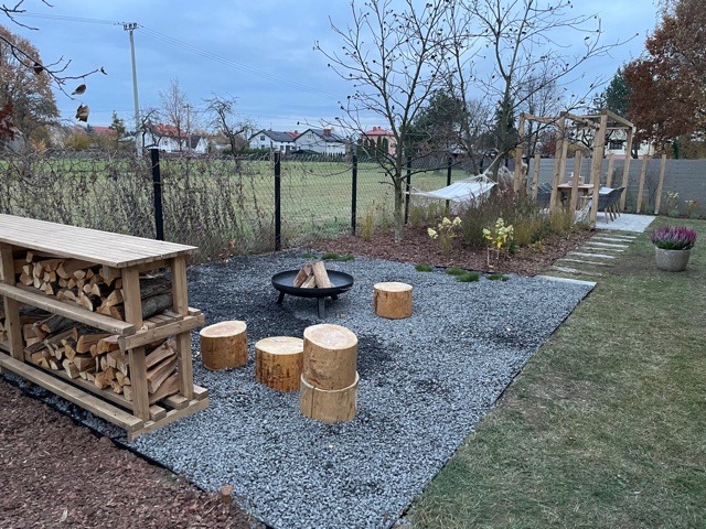 Ogród z miejscem na ognisko: palenisko otoczone drewnianymi siedziskami, drewniana wiata na drewno, hamak i pergola z meblami ogrodowymi, szara żwirowa nawierzchnia, widok na trawnik i domy w tle.