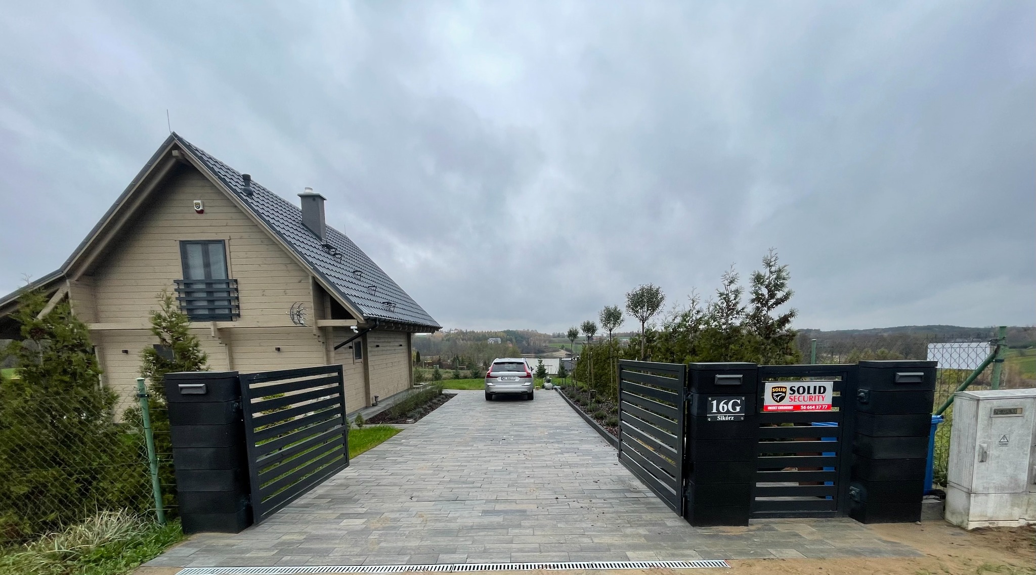 Nowoczesny dom jednorodzinny z drewnianą elewacją i szarą kostką brukową na podjeździe, widok zza otwartej, czarnej bramy wjazdowej, szare niebo w tle.