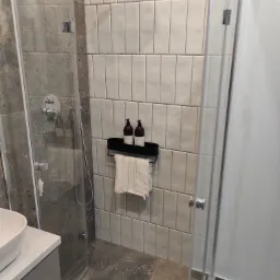 Nowoczesna kabina prysznicowa z deszczownicą, białe pionowe kafelki, szara ściana z mozaiką, półka z brązowymi butelkami i ręcznikiem.