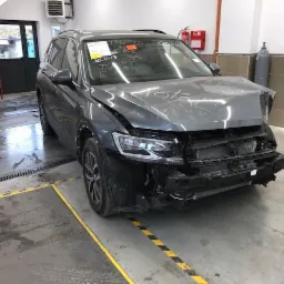 Uszkodzony, szary samochód osobowy marki Volkswagen Tiguan, z widocznymi wgnieceniami na masce i zderzaku, stoi w warsztacie blacharsko-lakierniczym przed naprawą. Na przedniej szybie widoczne...