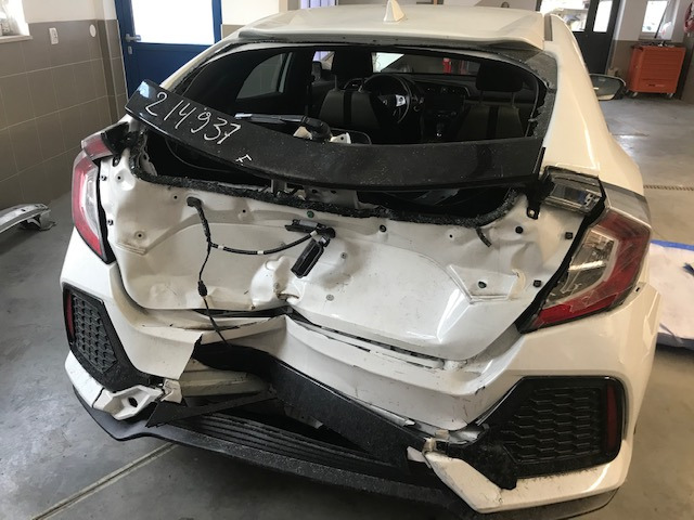 Biały samochód osobowy po kolizji, widoczne poważne uszkodzenia tylnej części pojazdu, w tym zgnieciony zderzak, uszkodzona klapa bagażnika i wybita tylna szyba. Tabliczka z numerem...