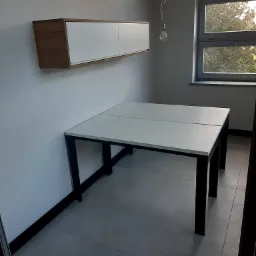 Biały blat biurka z czarnymi nogami, dopasowany do wiszącej szafki z drewnianymi bokami, w minimalistycznym wnętrzu z oknem i żarówką na kablu.