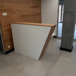 Nowoczesna lada recepcyjna z białym frontem i drewnianymi elementami wykończeniowymi, ustawiona w minimalistycznym wnętrzu z drewnianą ścianą w tle.