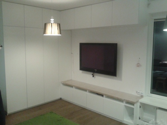 Biała zabudowa ściany z szafami i szafkami pod telewizorem, minimalistyczny styl, dywanik na podłodze.