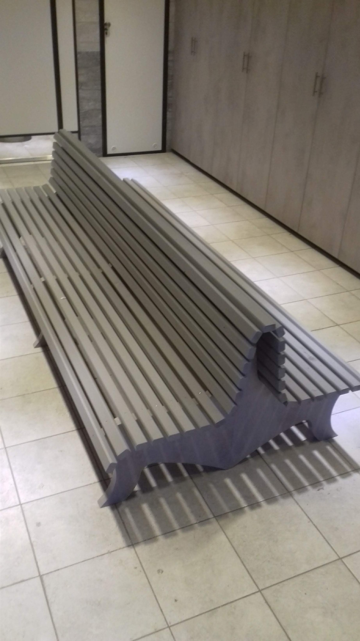 Szara ławka z drewnianych listew z oparciem, o nietypowej konstrukcji nóg, ustawiona na kafelkach w pomieszczeniu z szafami w tle.