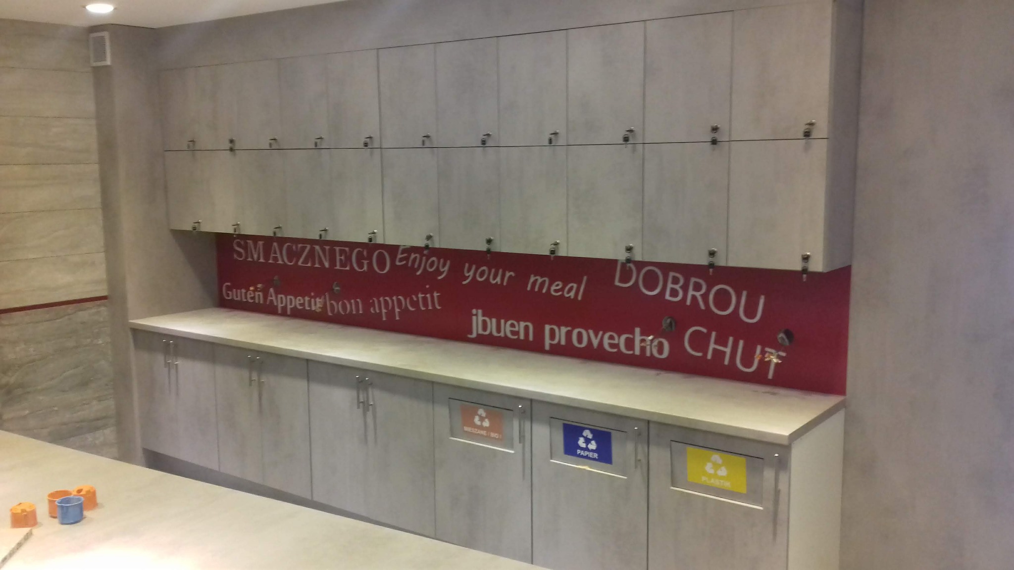 Szara zabudowa meblowa z szafkami zamykanymi na klucz, nad blatem z napisem 'Smacznego' w różnych językach i koszami do segregacji śmieci.