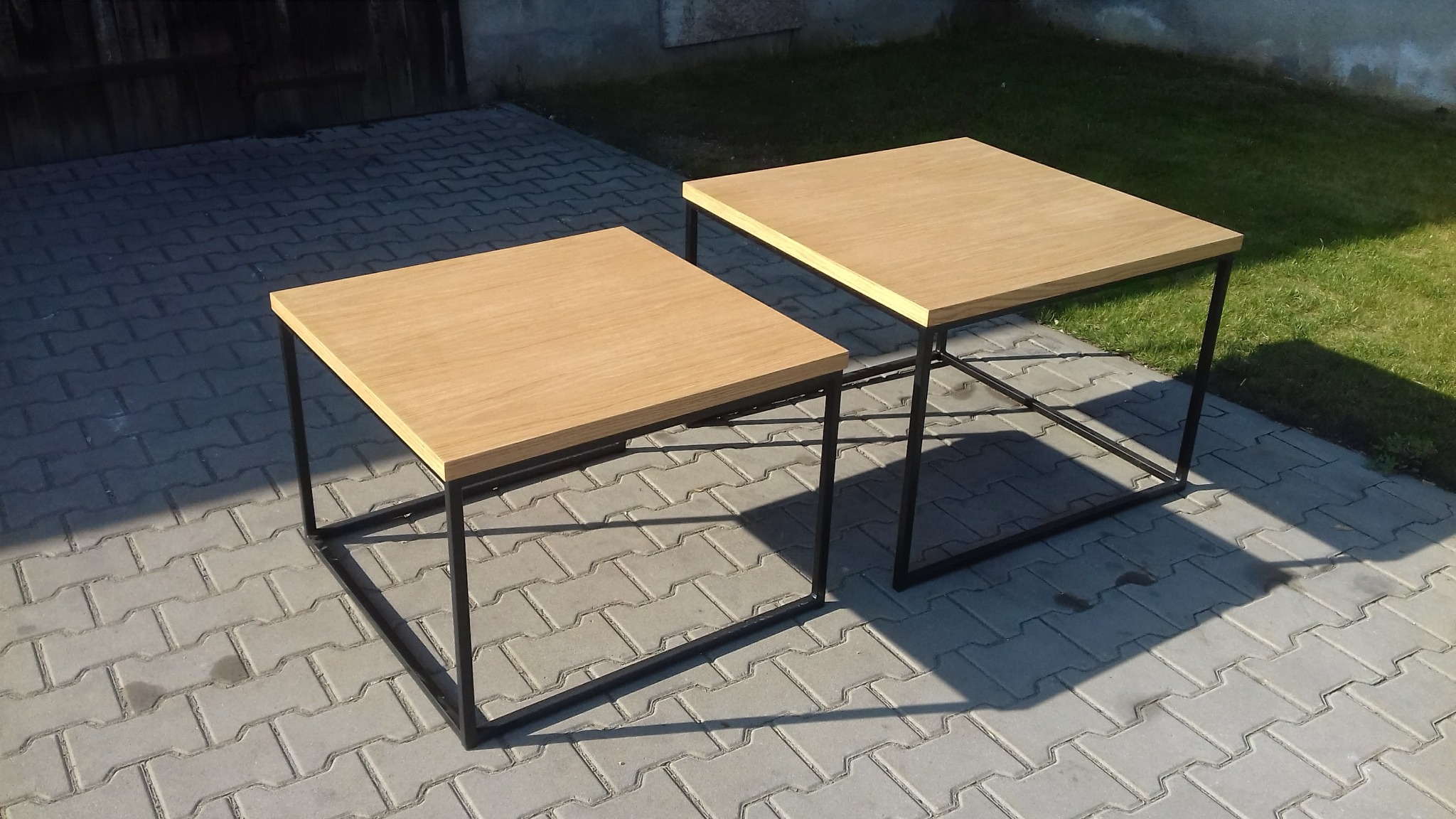 Dwa identyczne, kwadratowe stoliki kawowe z jasnego drewna na czarnych, metalowych nogach, ustawione na szarej kostce brukowej w ogrodzie.