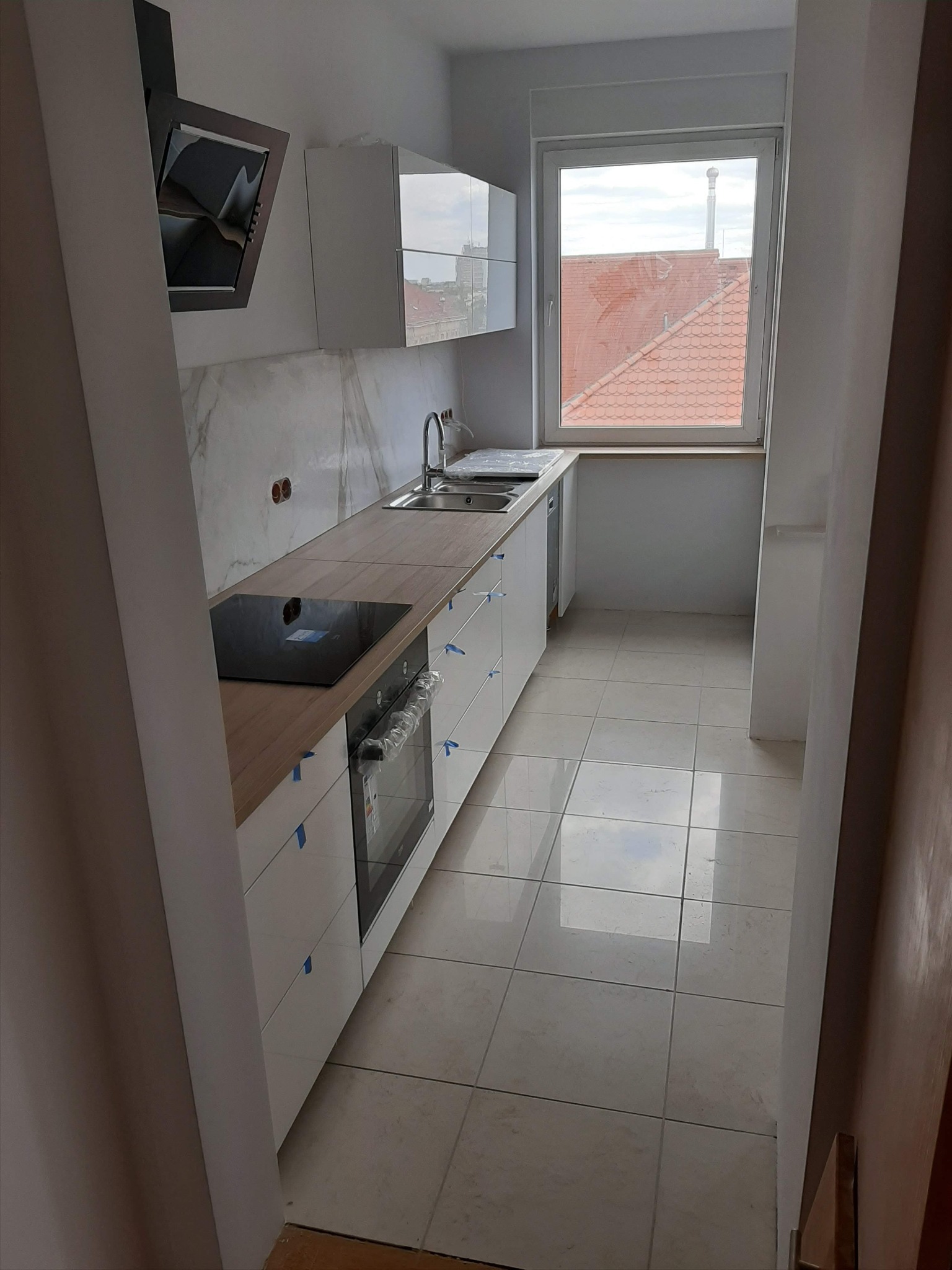 Wąska kuchnia w trakcie montażu, widoczne białe szafki z drewnianym blatem, płyta indukcyjna, piekarnik w folii ochronnej i marmurowe panele ścienne.