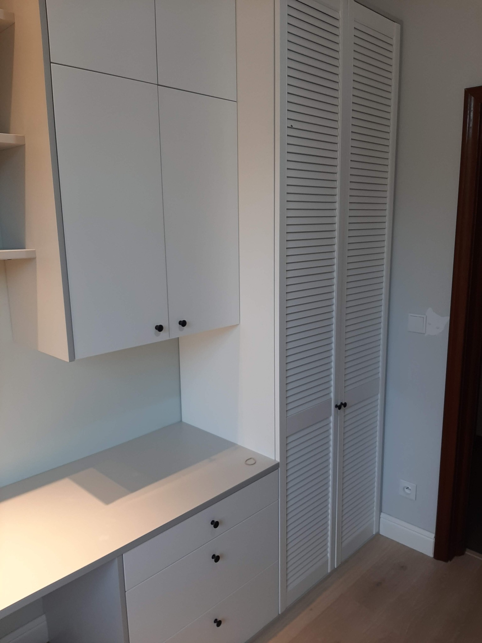 Biała szafa wnękowa z lamelowymi drzwiami, połączona z biurkiem i szafkami wiszącymi w jasnym wnętrzu w Poznaniu.